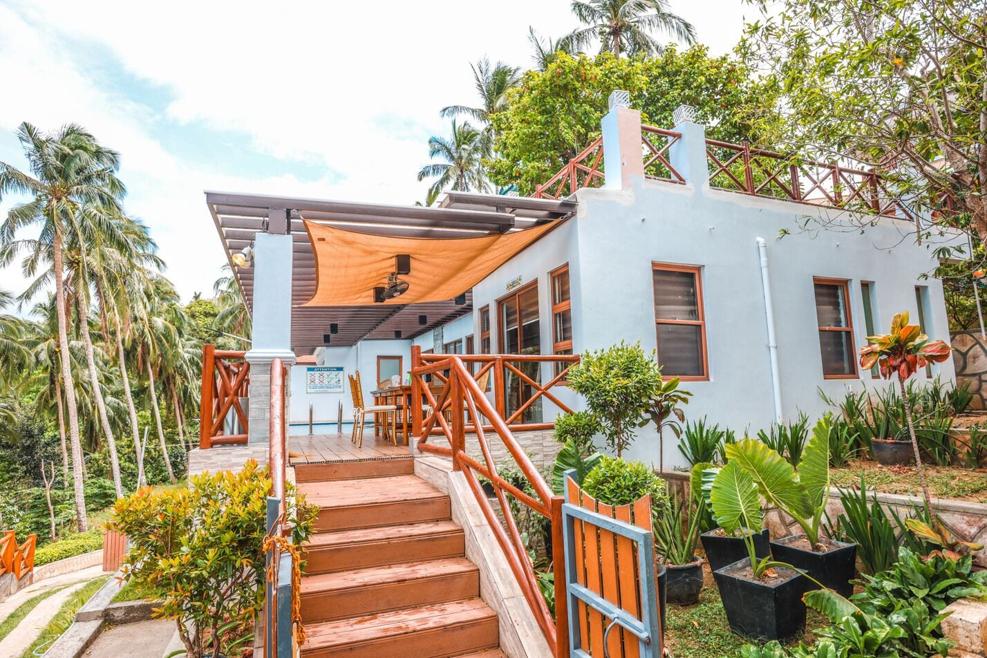A two-story building with a staircase leading to a covered outdoor area, surrounded by tropical vegetation.
