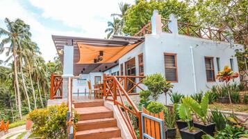 A two-story building with a staircase leading to a covered outdoor area, surrounded by tropical vegetation.