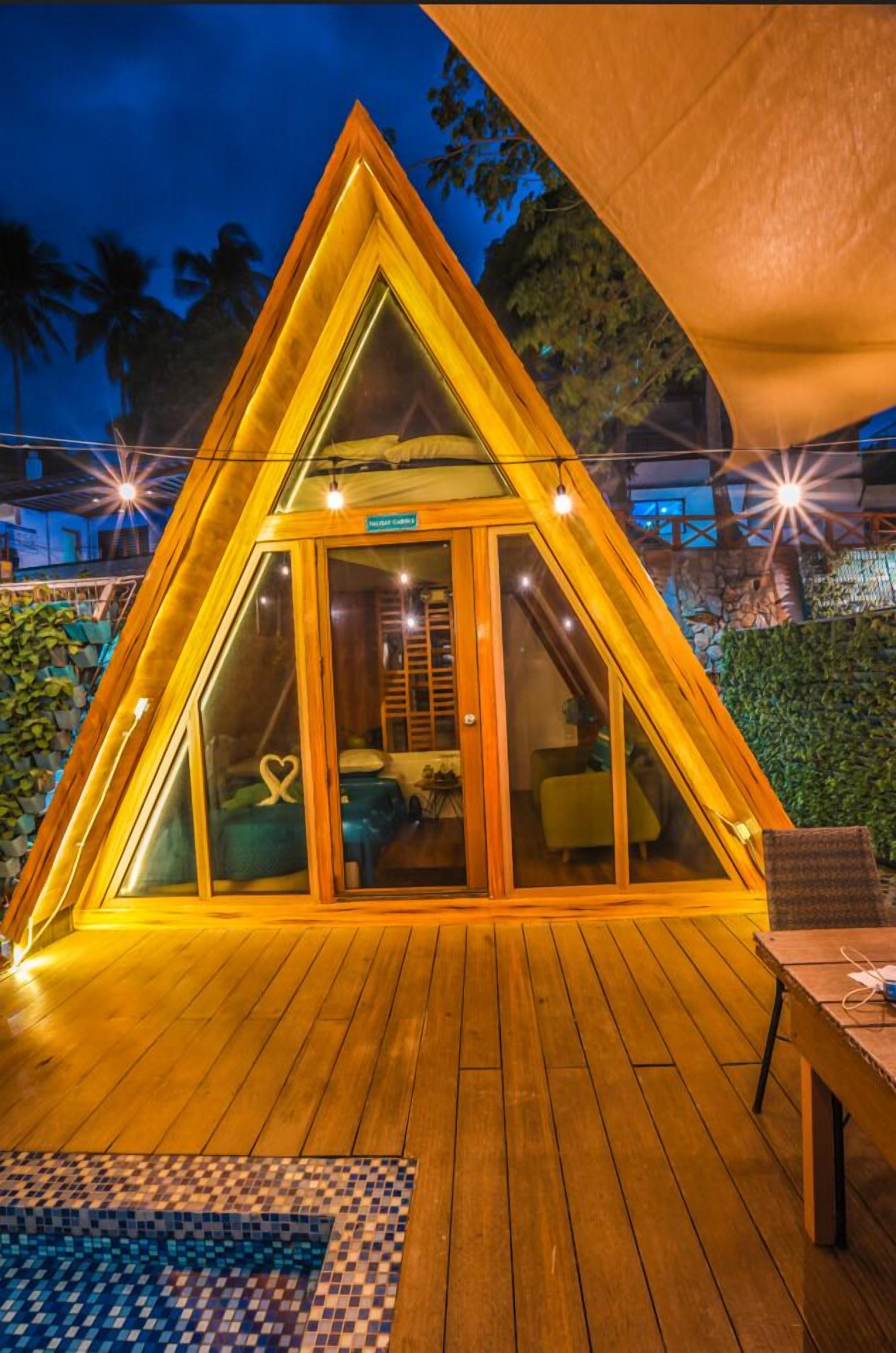 Un bâtiment de forme triangulaire doté d’une entrée avec une porte en verre, d’une terrasse en bois et d’un espace extérieur aménagé pour s’asseoir.