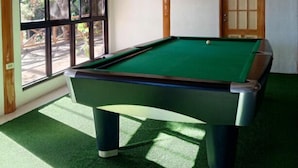 Una mesa de billar en una habitación con techo de madera, ventanas amplias y una puerta.