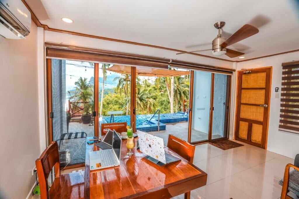 A room with a wooden dining table, chairs, and a laptop. There is a ceiling fan and a view of a pool and palm trees outside.