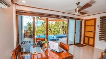 A room with a wooden dining table, chairs, and a laptop. There is a ceiling fan and a view of a pool and palm trees outside.