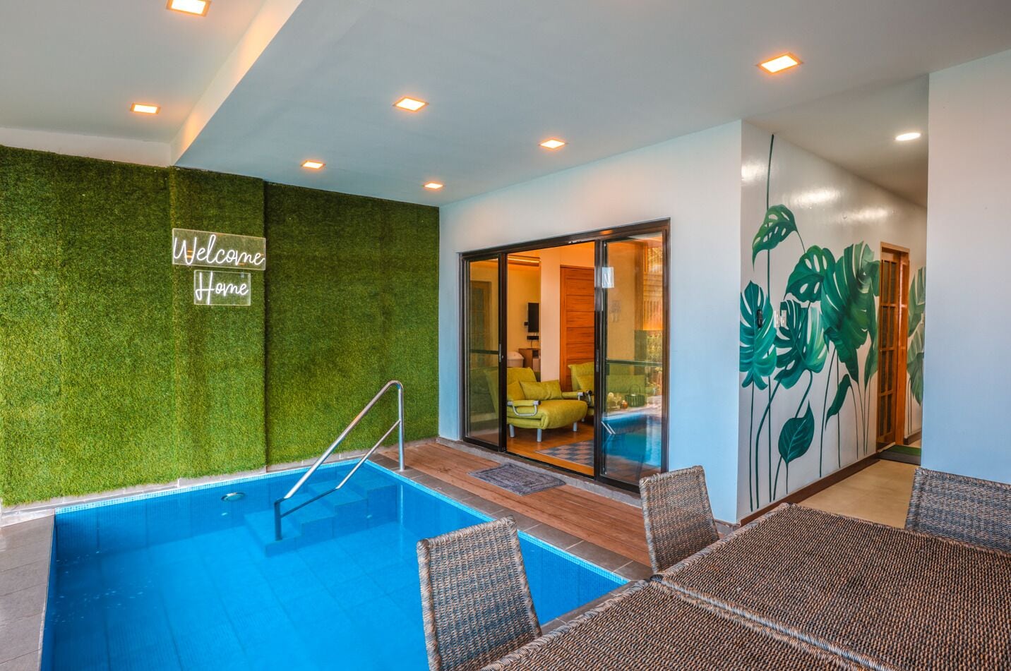A swimming pool with a glass enclosure, a 'Welcome Home' sign, and a room with a green wall and tropical leaf mural.