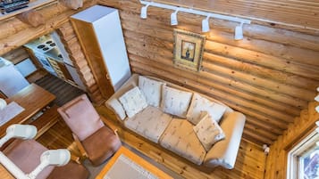 Interior de cabana com piso de madeira, um sofá com almofadas florais, uma mesa de jantar com cadeiras, uma área de cozinha e um lustre no teto.