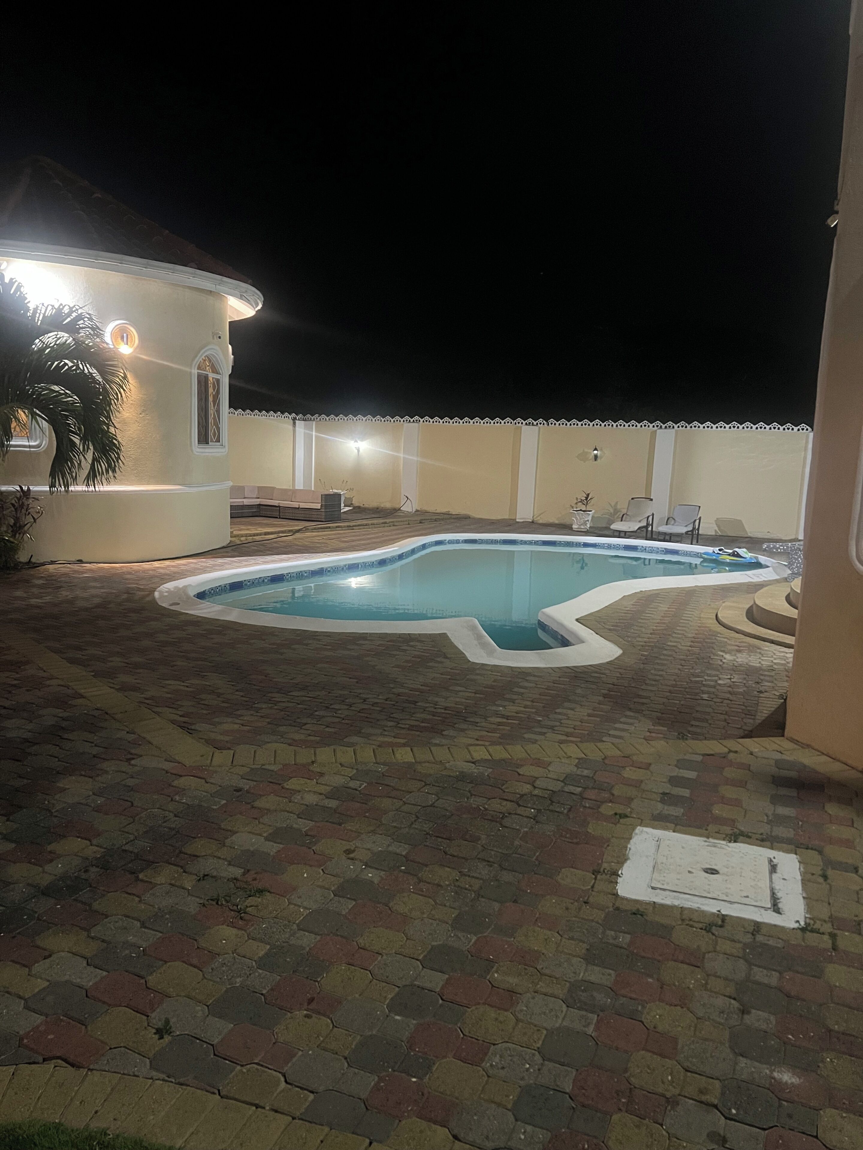A swimming pool with a brick patio, a small building with a light, and a white fence.