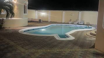 A swimming pool with a brick patio, a small building with a light, and a white fence.