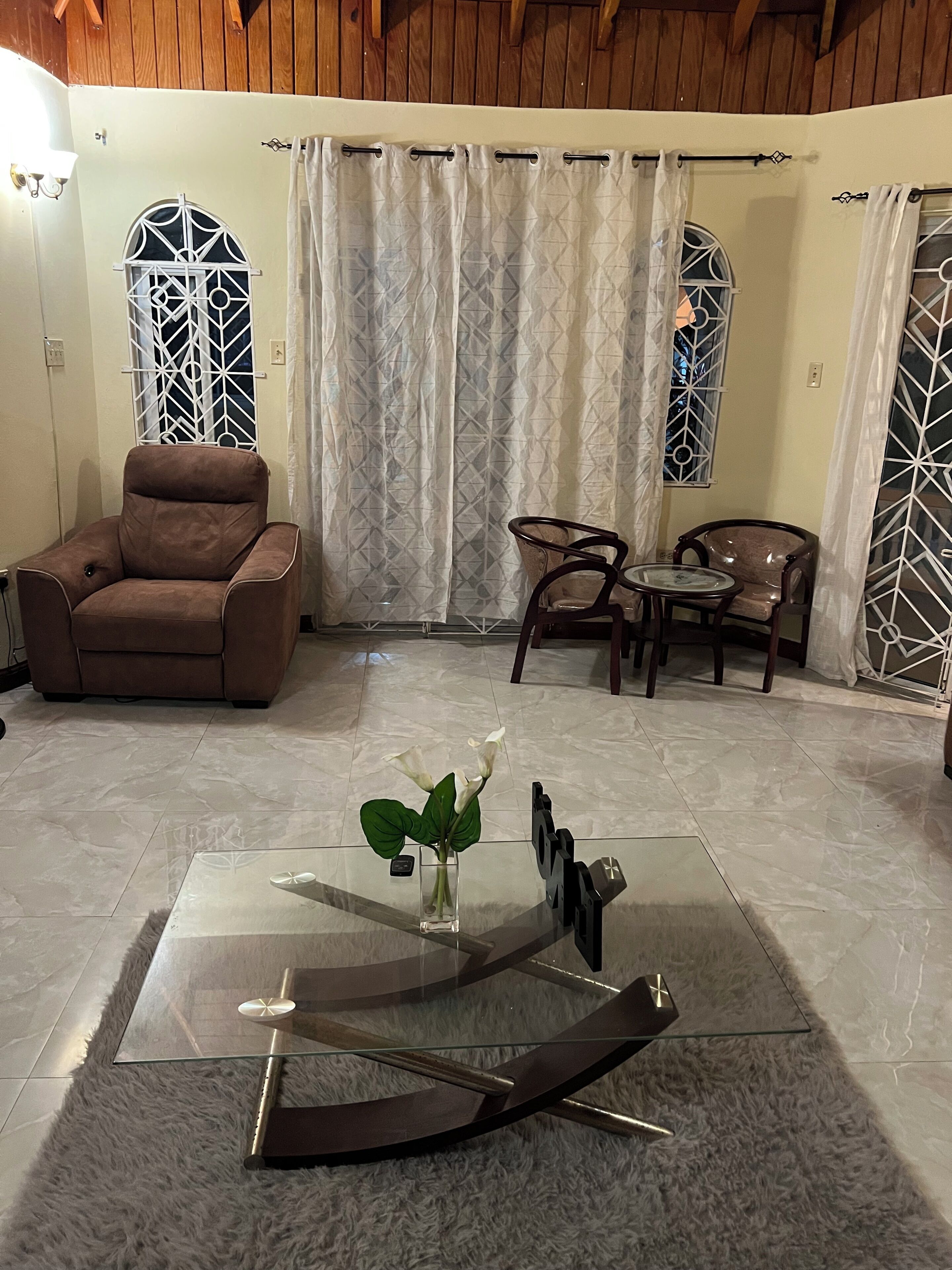 A living room with a brown leather chair, a glass coffee table with a vase of flowers, and a patterned curtain.