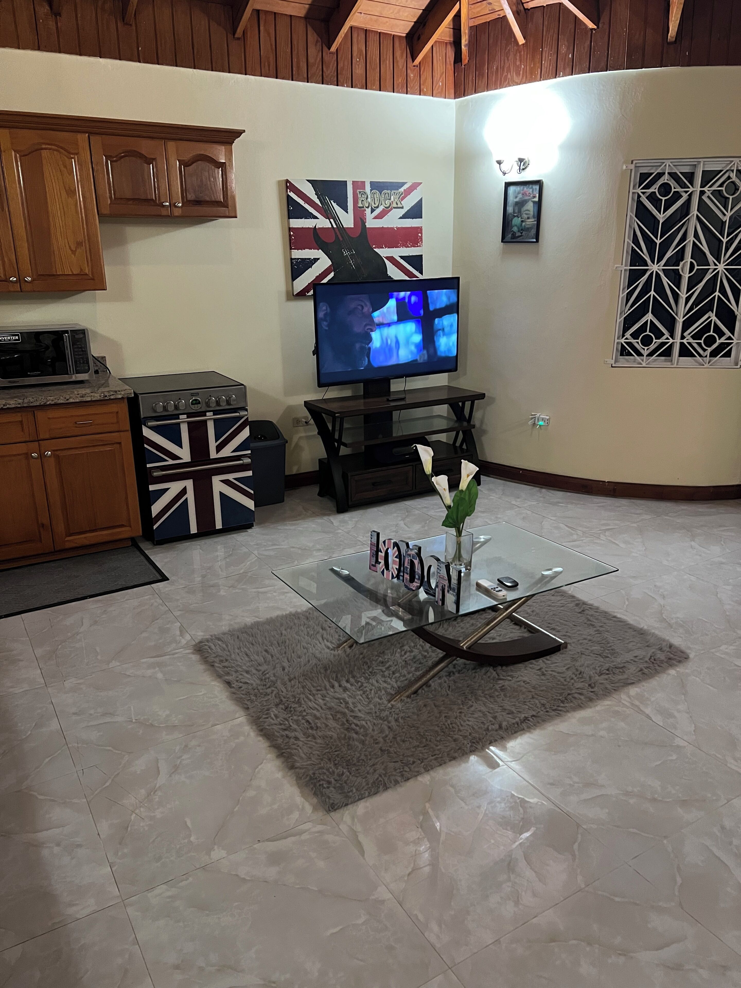 Een woonkamer met een televisie, houten kasten, een oven met Union Jack thema en een glazen bijzettafeltje met een vaas.