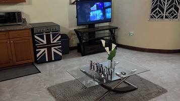 Een woonkamer met een televisie, houten kasten, een oven met Union Jack thema en een glazen bijzettafeltje met een vaas.