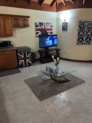 Una sala de estar con televisor, armarios de madera, un horno con temática de la Union Jack y una mesa de centro de vidrio con un jarrón.