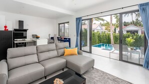 A modern living room with a grey sectional sofa, a flat-screen TV, a dining area with a blue table, and a large sliding glass door leading to a pool. - Buddha Bliss Luxury Villa (Tambon Ko Pha-Ngan)
