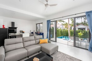 A modern living room with a grey sectional sofa, a flat-screen TV, a dining area with a blue table, and a large sliding glass door leading to a pool. - Buddha Bliss Luxury Villa (Tambon Ko Pha-Ngan)