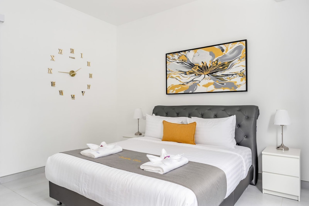 A hotel room with a bed, a nightstand, a wall clock, and a piece of abstract art.