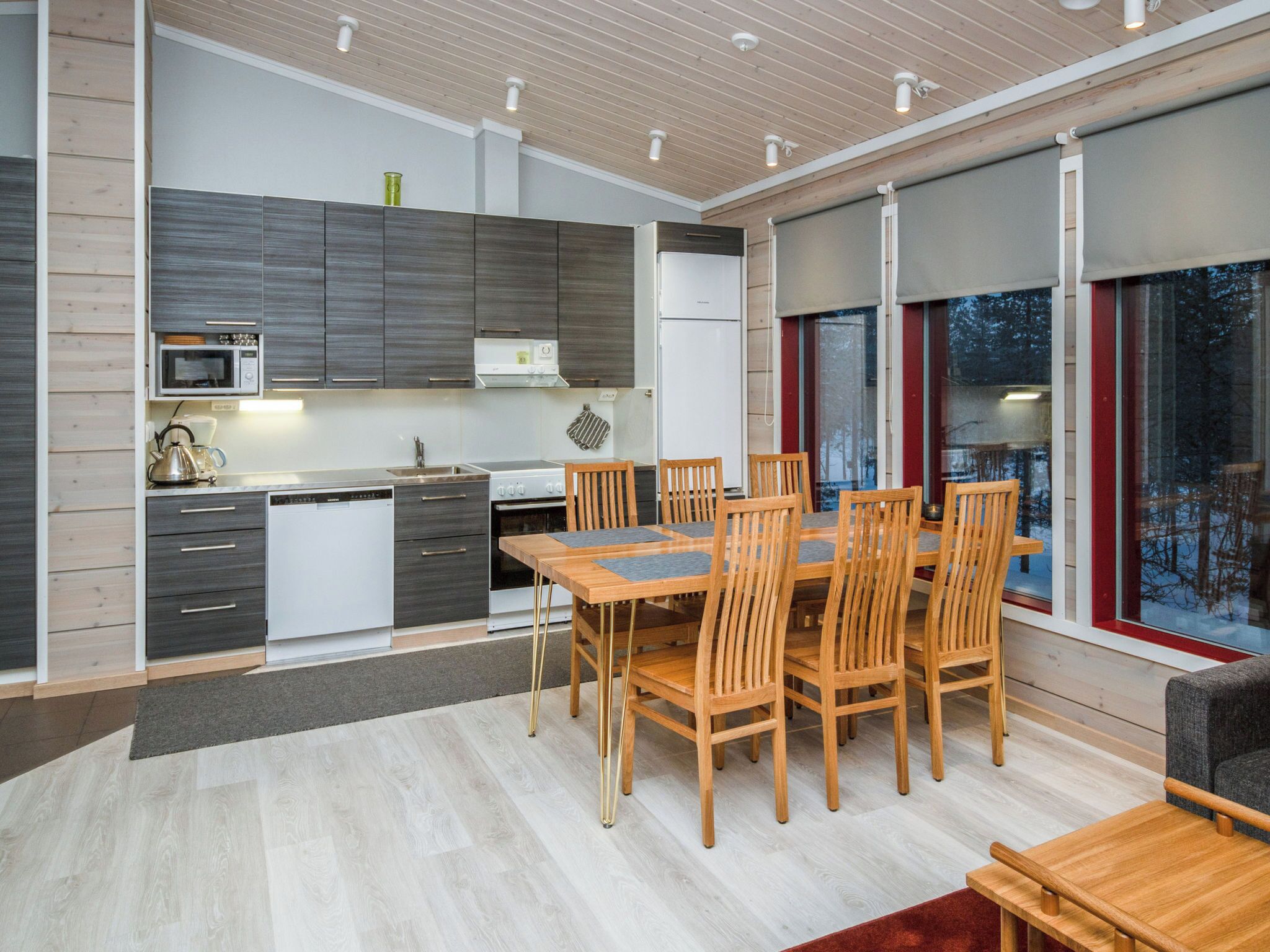 A modern kitchen with a dining area, featuring a large wooden table and chairs, a built-in oven, and a view through large windows.