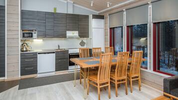 A modern kitchen with a dining area, featuring a large wooden table and chairs, a built-in oven, and a view through large windows.