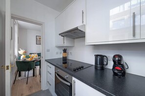 A modern kitchen with white cabinets, a black countertop, a built-in stove, and a coffee maker. - Bright Central 1BR 1BA steps from Oxford Street (London)