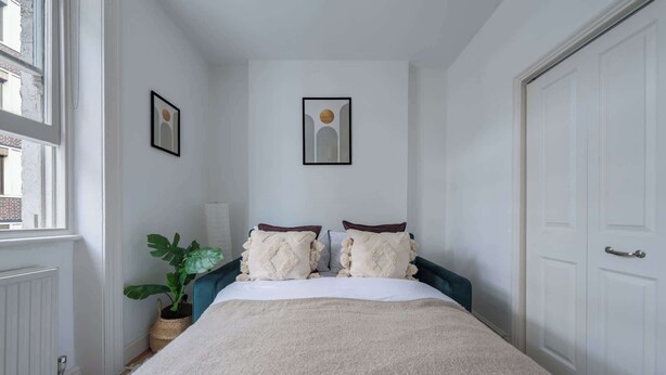 Een slaapkamer met een bed, twee kussens, een kamerplant en een lijst aan de muur.