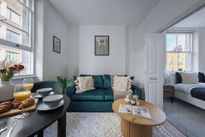 A modern living room with a teal sofa, a wooden coffee table, and a dining area with breakfast items. There are large windows providing natural light and a view of the outside. - Bright Central 1BR 1BA steps from Oxford Street (London)