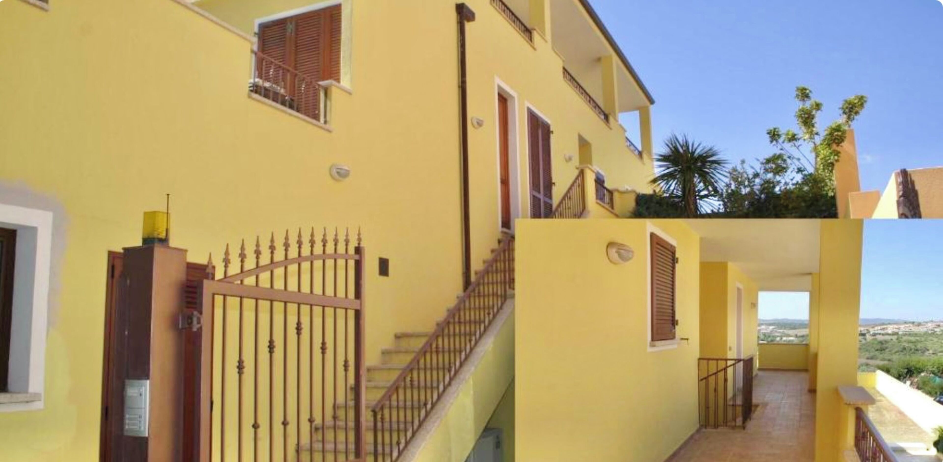 A yellow building with a gated entrance, a staircase, and a balcony with a view of the landscape.