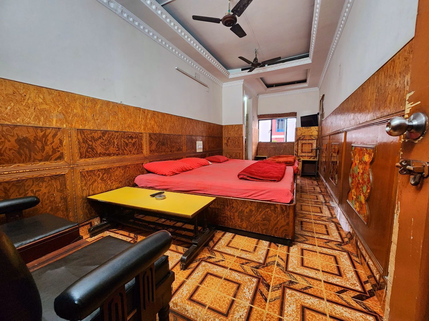 A compact hotel room with a bed, a small table, a chair, a ceiling fan, and a patterned floor.
