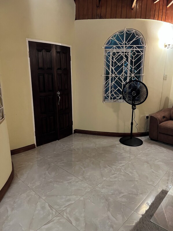 A room with a tiled floor, a standing fan, a door, and a window with a decorative grille.