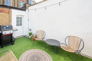 Eine Terrasse mit Grill, zwei Korbstühlen, einem runden Tisch und Kübelpflanzen.