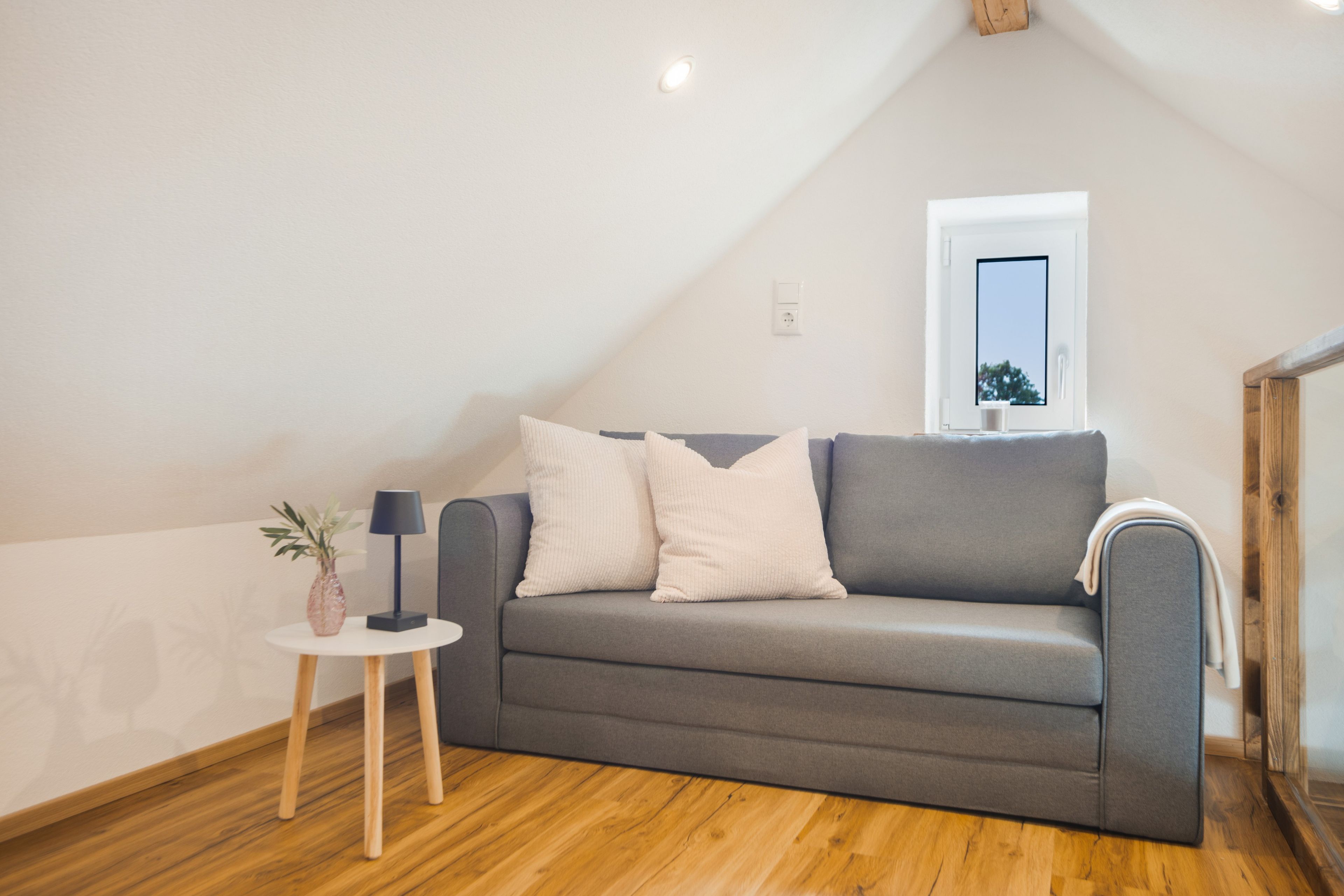 Ein modernes Wohnzimmer mit einem grauen Sofa, einem kleinen Beistelltisch mit Lampe und einem Holzboden.