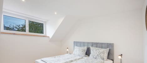 A bedroom with a sloped ceiling, a bed with patterned bedding, two bedside lamps, and a window with a view of trees.