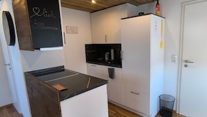 Ett kompakt kök med vita skåp, en svart bänkskiva och en liten köksö. Det finns en svart tavla där det står ”Willkommen”, och en skylt som lyder ”Partys Küche”.