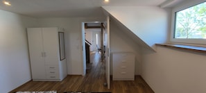 Una habitación con piso de madera, un armario blanco, un escritorio con cajones y una ventana con vista a árboles.