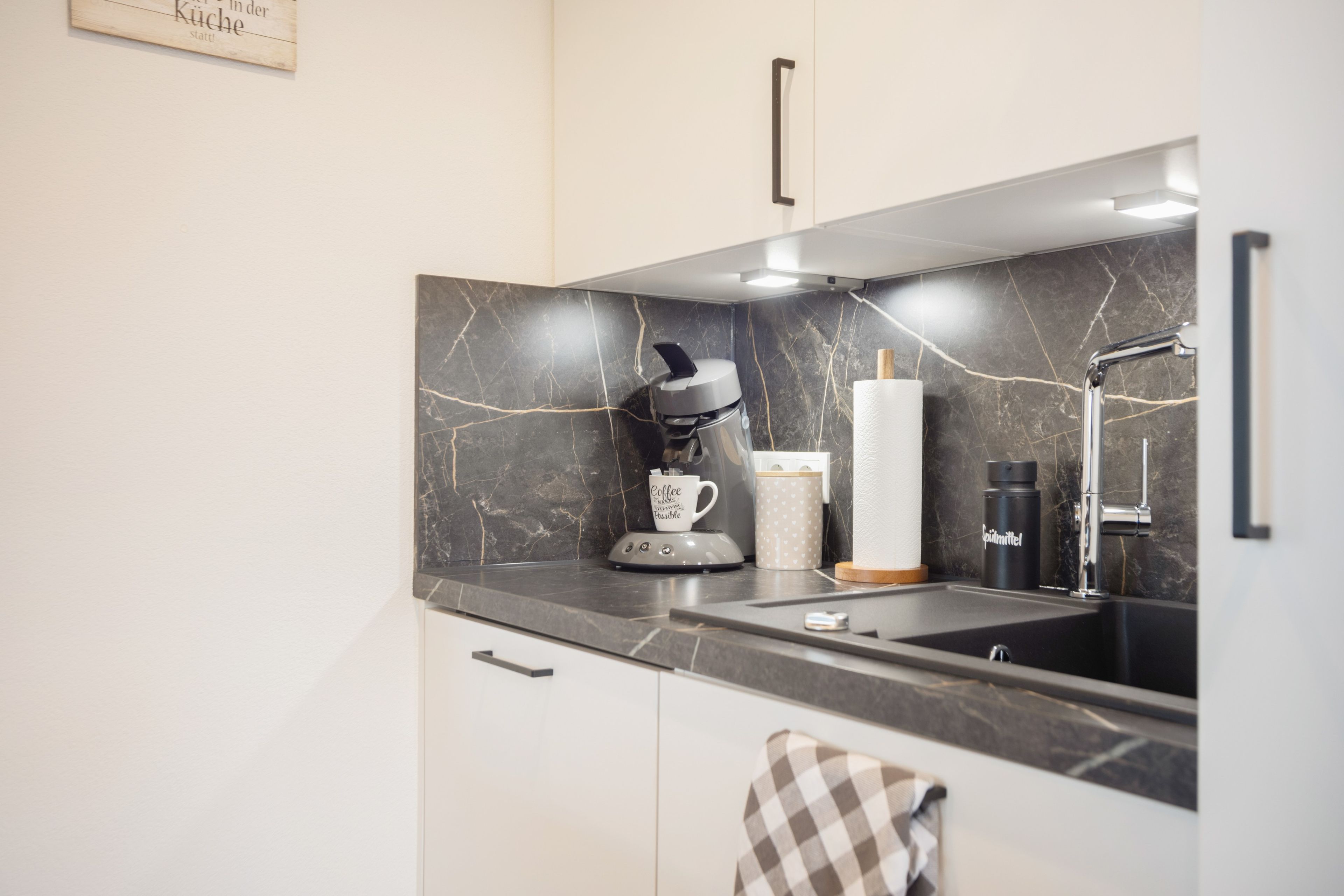 Ett modernt kök med en svart marmorvägg bakom diskbänken, en kaffebryggare, en vattenkokare och en diskho.