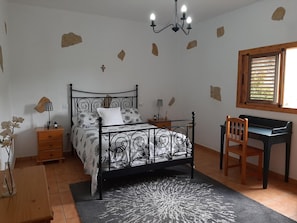 A bedroom with a bed, bedside tables, a desk, a chair, a window with blinds, and a chandelier.