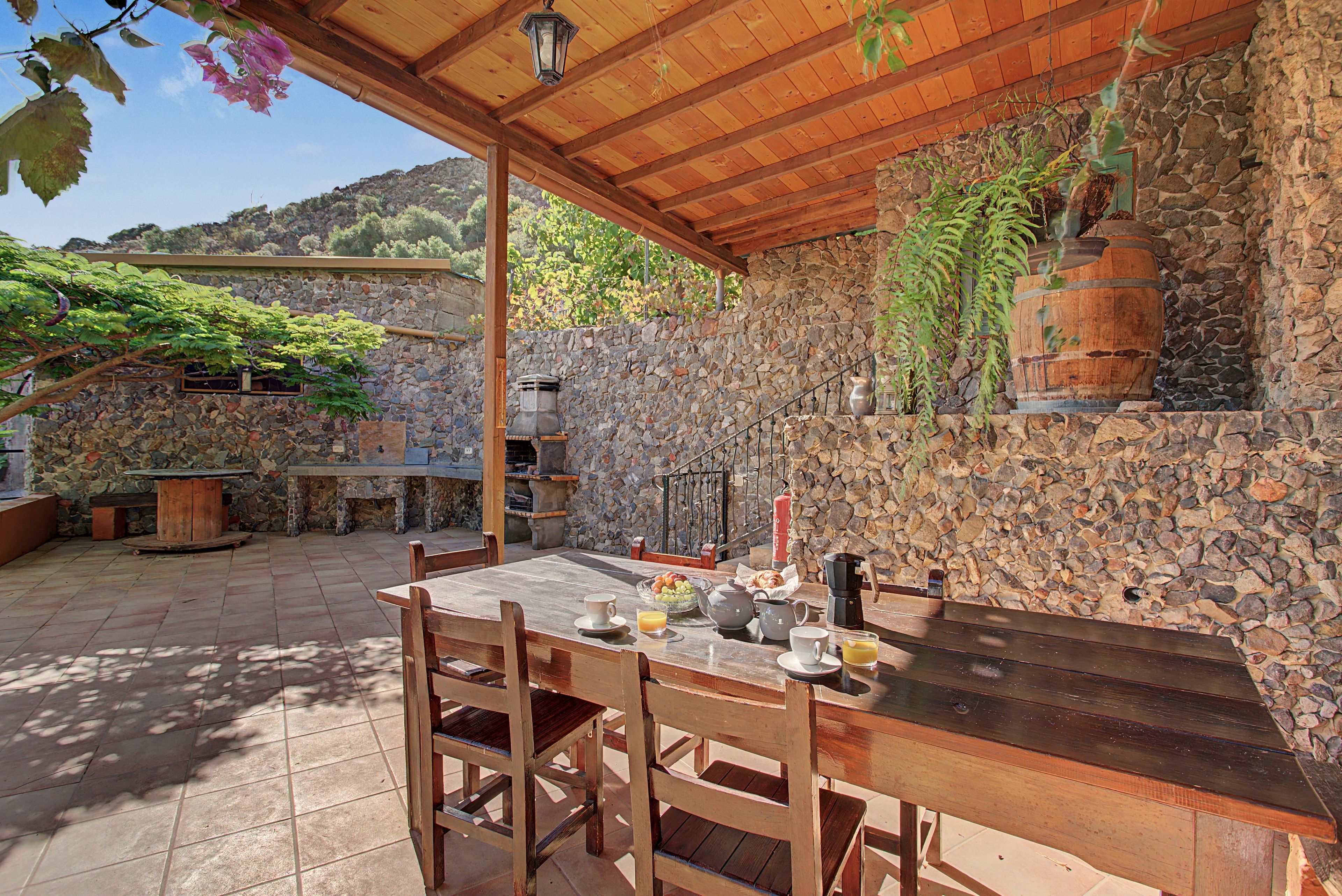 Un patio exterior con pared de piedra, mobiliario de madera y chimenea de piedra.