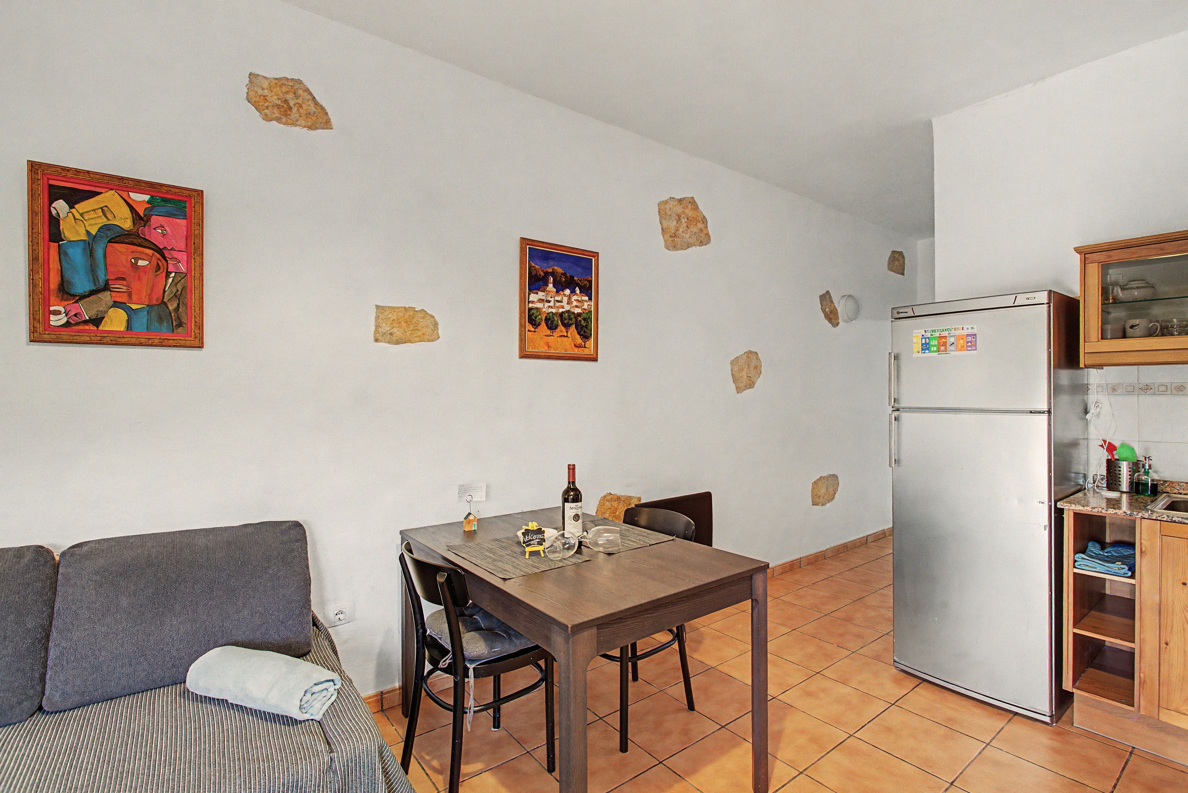 Un espacio habitable compacto con una mesa de comedor, sillas, un sofá, una zona de cocina con refrigerador y dos cuadros colgados en la pared.