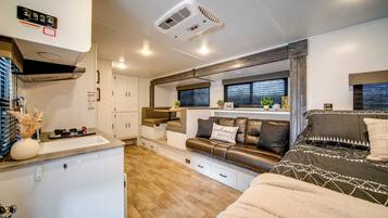 A compact RV interior with a kitchenette, a seating area with a sofa and a bed, and a window with blinds.