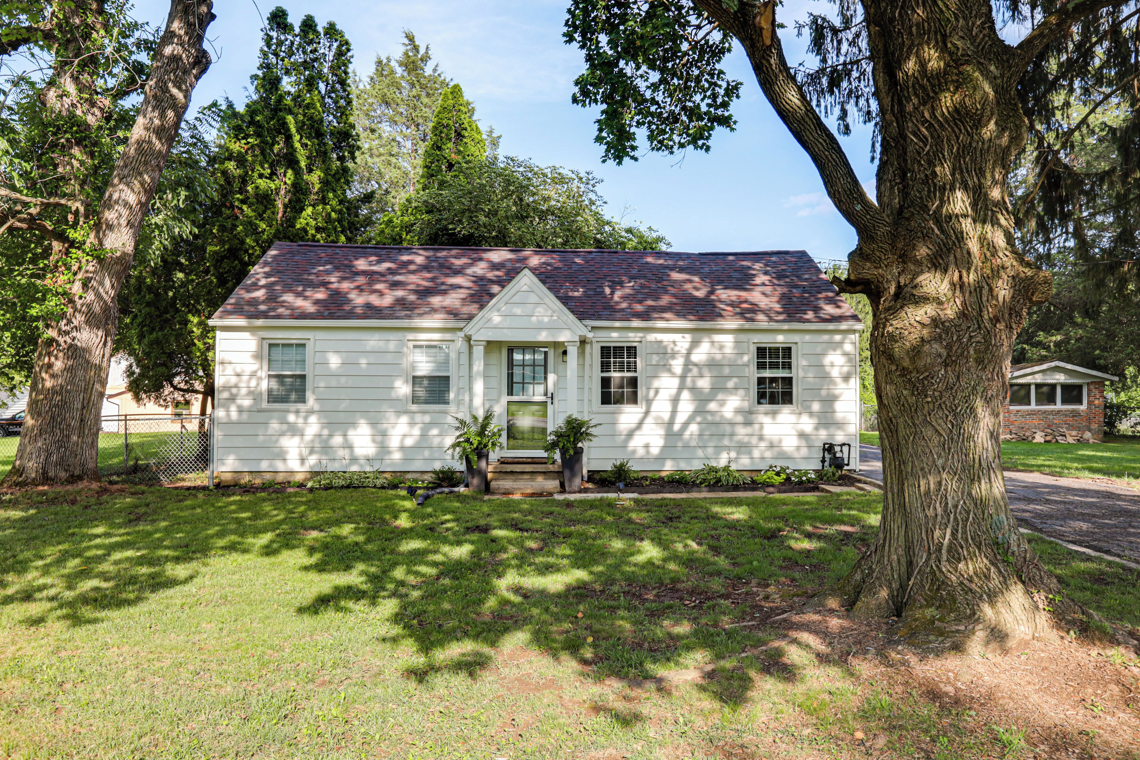 A single-story house with a gabled roof, a front porch with a bench, and a small front yard with a tree.