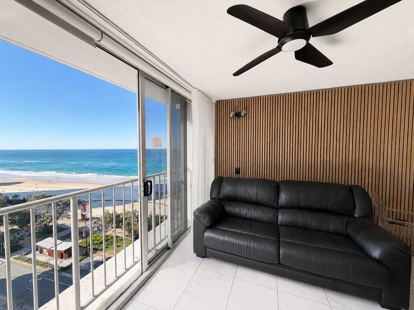 Un balcón con vista a la playa, un sofá de cuero negro y un ventilador de techo.