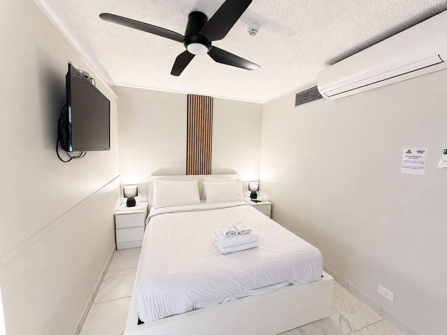 Quarto de hotel com cama, ventoinha no teto, televisão fixada na parede, abajures e um aviso na parede.