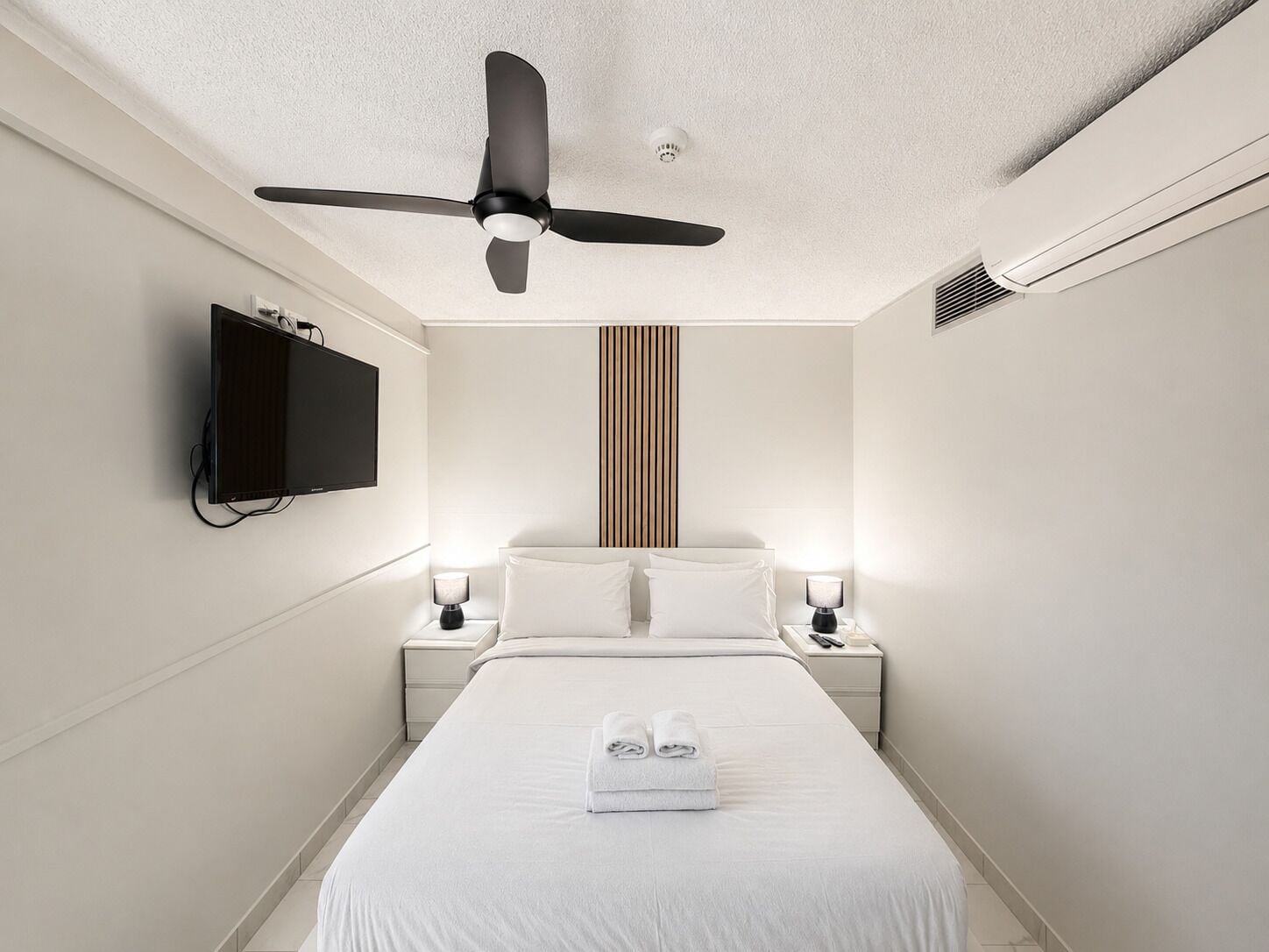 Habitación de hotel con cama, ventilador de techo, televisor montado en la pared y lámparas de noche.