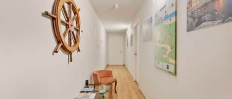 A hallway with a wooden floor, a table with magazines, a chair, a ship's wheel decoration, and a welcome poster.