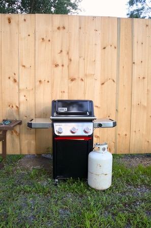 A portable gas grill with a propane tank, positioned outdoors against a wooden fence.