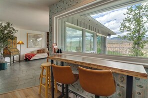 A cozy room with a large window, a wooden bar counter, two bar stools, a sofa with a patterned throw, a lamp, and a framed picture on the wall.