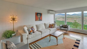 Ein modernes Wohnzimmer mit einem weißen Sofa, einem Couchtisch und Blick auf eine gebirgige Landschaft durch große Fenster.
