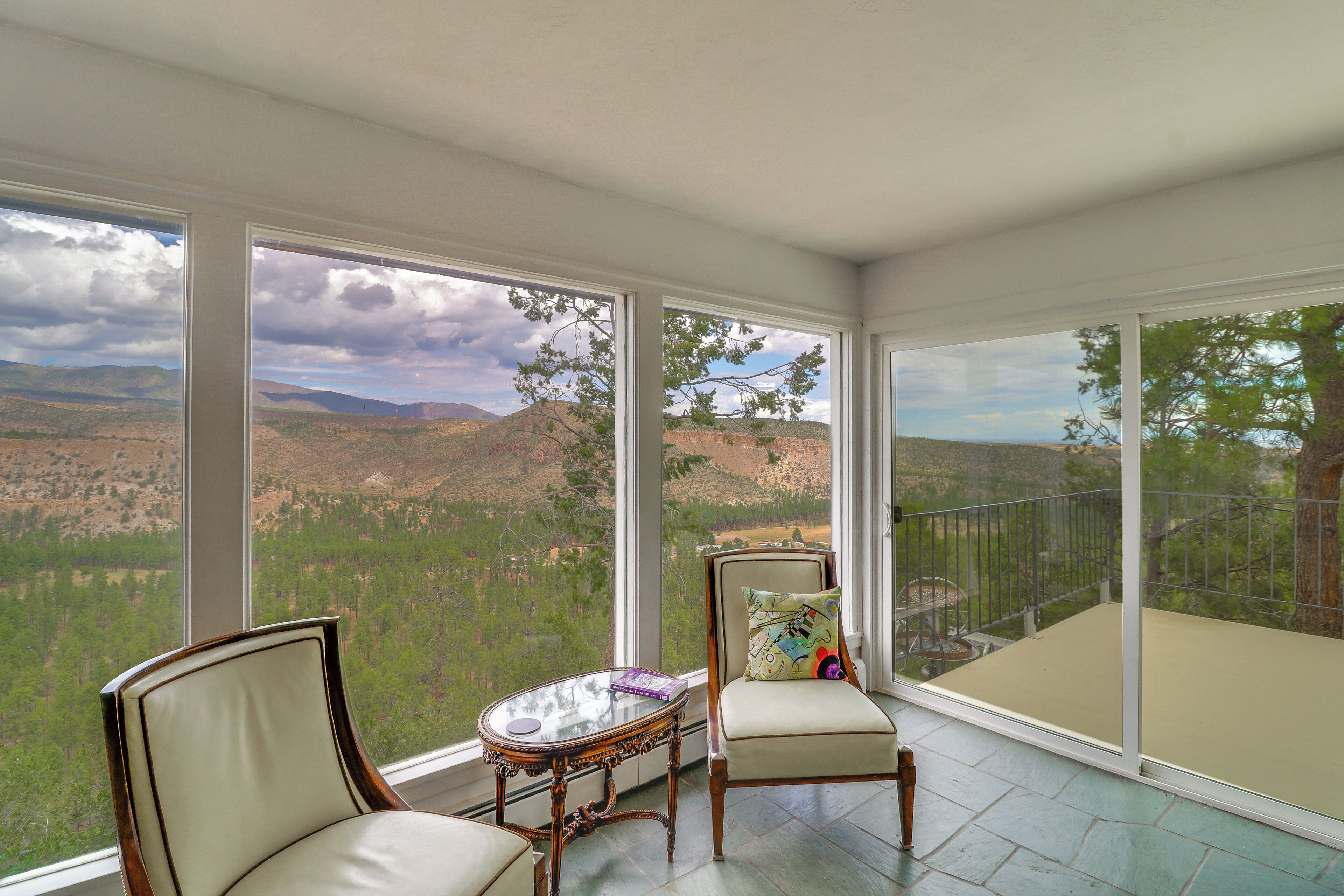 A room with large windows offering a view of a mountainous landscape. There are two chairs and a small table with a decorative pillow.