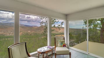A room with large windows offering a view of a mountainous landscape. There are two chairs and a small table with a decorative pillow.