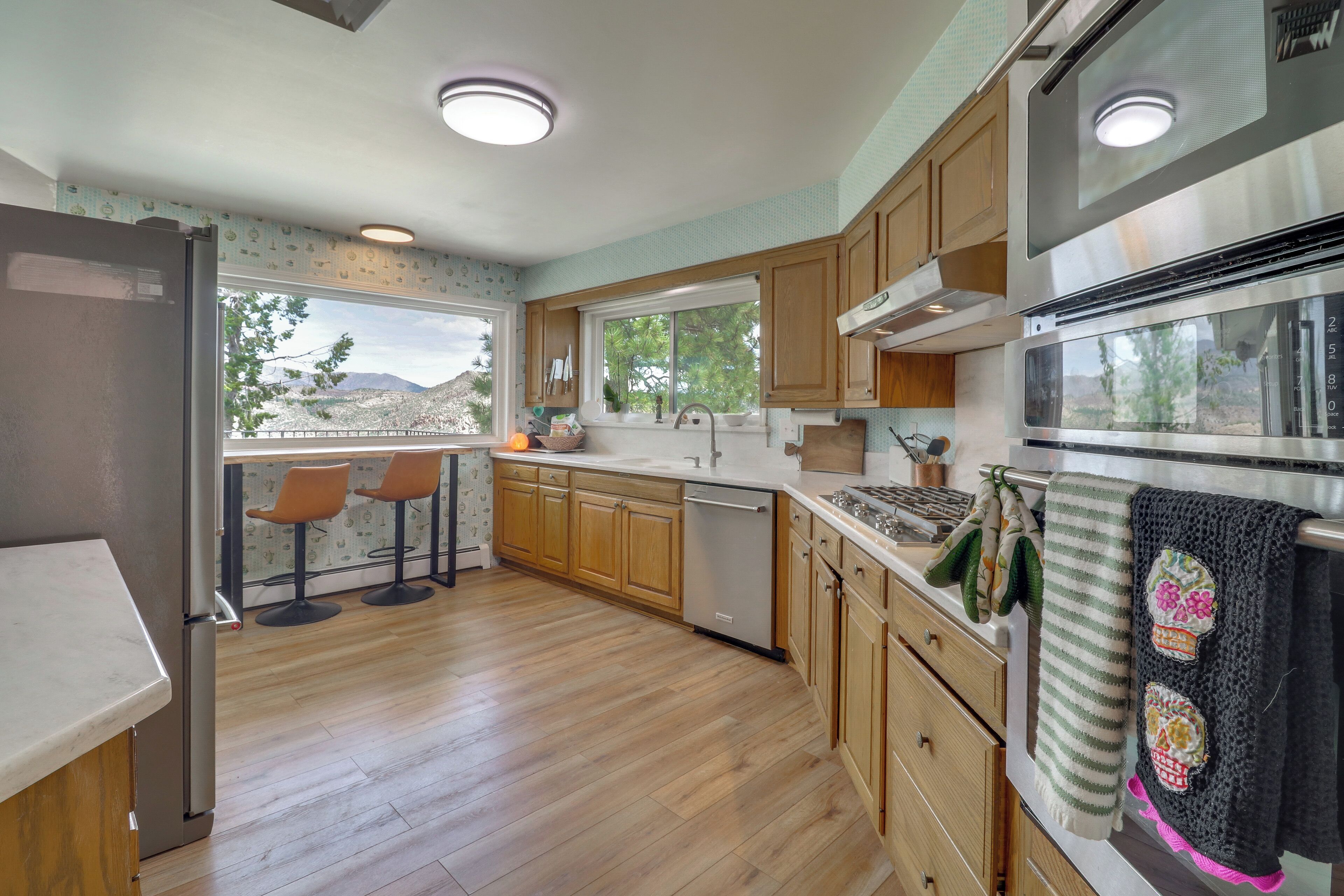 A kitchen with wooden cabinets, a stainless steel refrigerator, a microwave, and a window providing a view of trees and mountains.