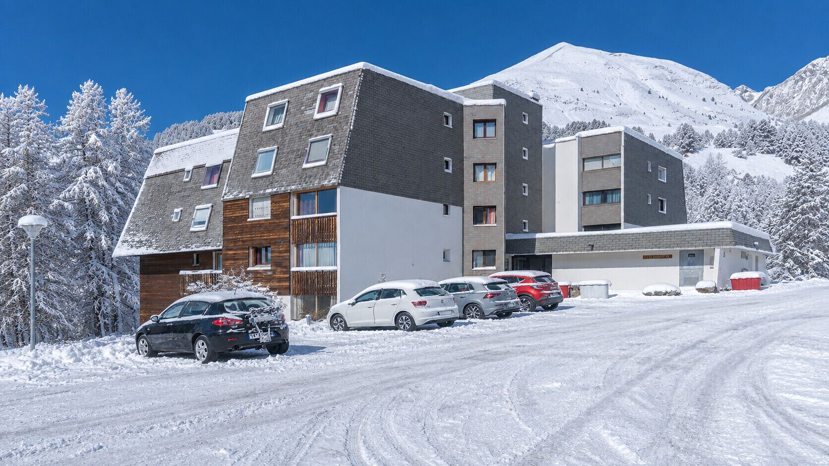A multi-story building with a parking area in front, surrounded by snow-covered trees and a mountain in the background.