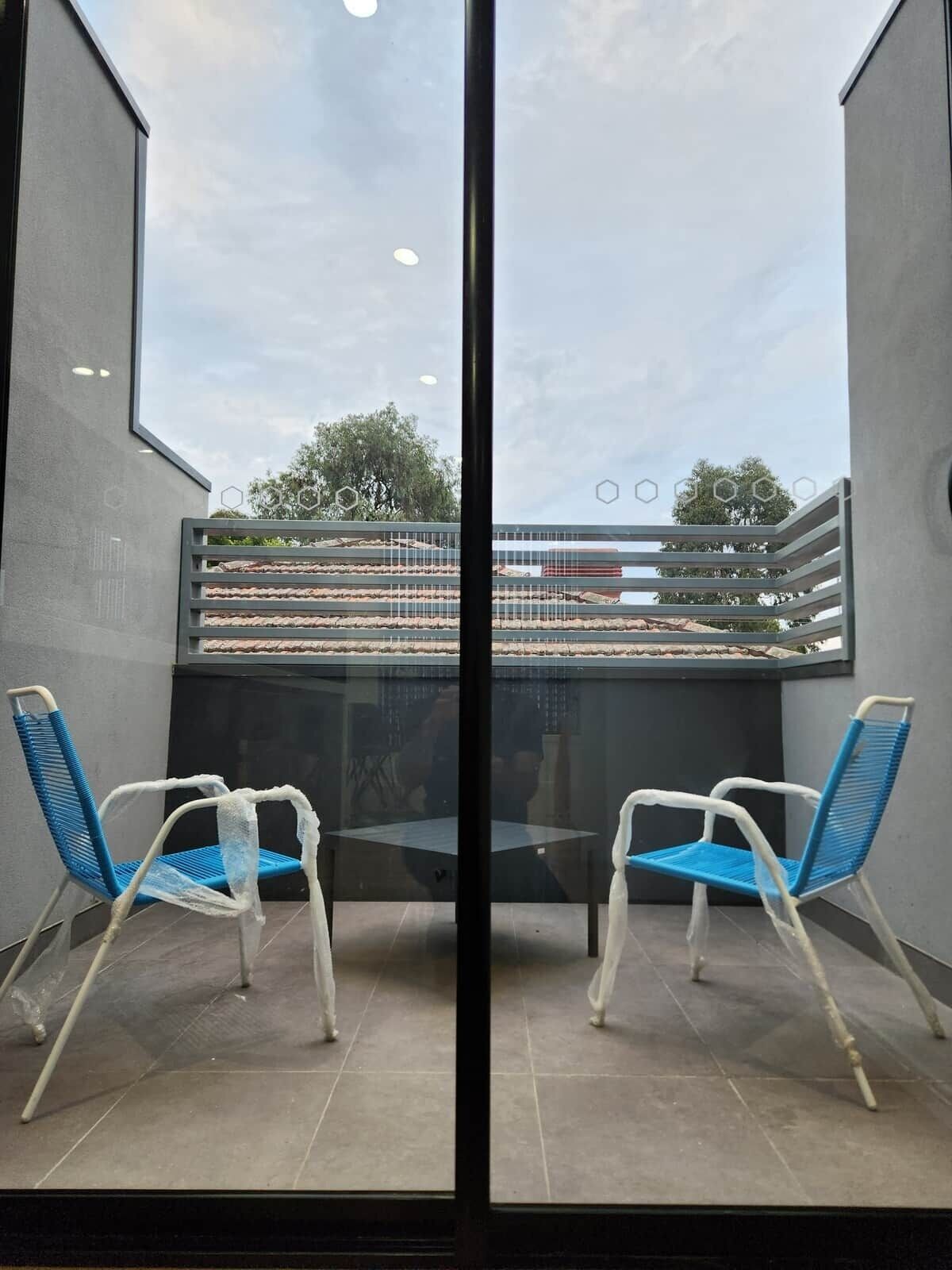 A room with a glass partition, two chairs, a table, and a view of a balcony with a railing and some greenery.