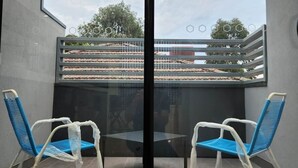 Ein Zimmer mit Glastrennwand, zwei Stühle, einem Tisch und Blick auf einen Balkon mit Geländer und etwas Grün.