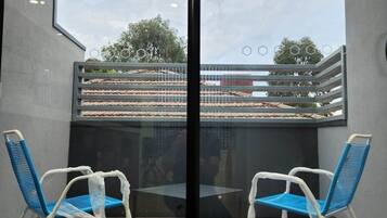 A room with a glass partition, two chairs, a table, and a view of a balcony with a railing and some greenery.